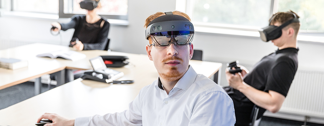 Ein Mann trägt eine AR-Brille und sitzt an einem Tisch, in der Hand bewegt er eine Computer-Maus, im Hintergrund sitzen zwei weitere Personen, die VR-Brillen tragen