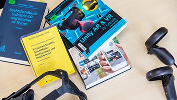 Vier Bücher liegen auf einem Tisch, daneben zwei Konsolen für eine VR-Brille