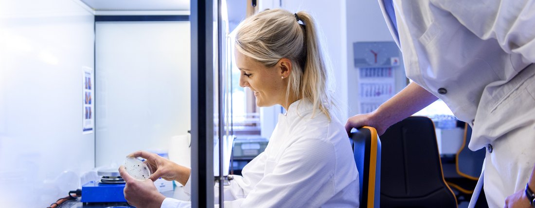 Zwei Mitarbeiter im Labor, eine Mitarbeiterin sitzend an einem Laborgerät, ein Mitarbeiter über die Schulter blickend