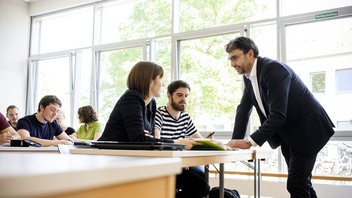 Studierende sitzen in einer Vorlesung und Professor steht am Tisch vor zwei Studierenden. Sie sprechen miteinander
