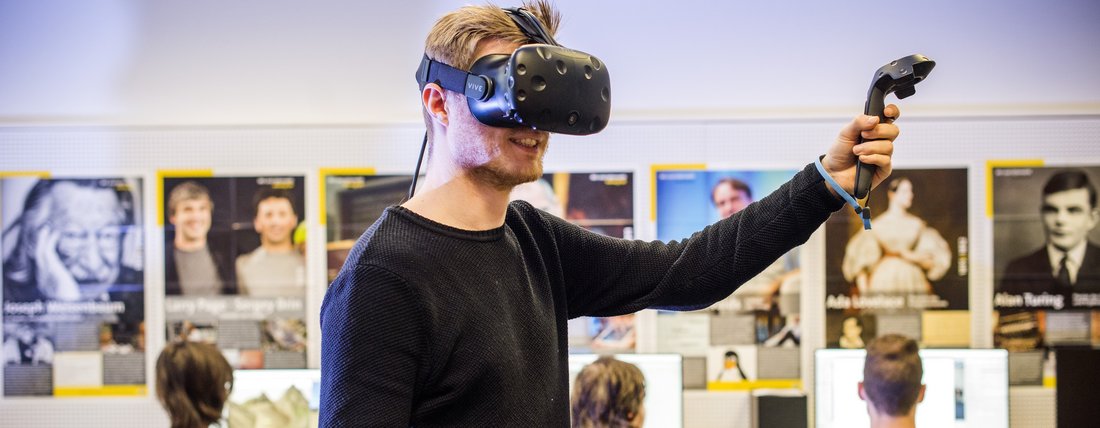 Ein blonder Mann mit einer Virtual Reality-Brille hält in seiner linken Hand eine Fernbedienung. Im Hintergrund sitzen drei Personen vor einer Wannd, mit dem Rücken gedreht zu dem Mann. An der Wand hängen viele Poster.