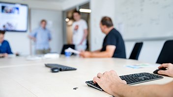 Man erkennt Hände, die auf einer Tastatur abgelegt sind, die Person sitzt an einem langen Meetingtisch, im Hintergrund sitzt eine weitere Person an diesem Tisch, drei andere Personen stehen am Ende des Raumes nah am Tisch 