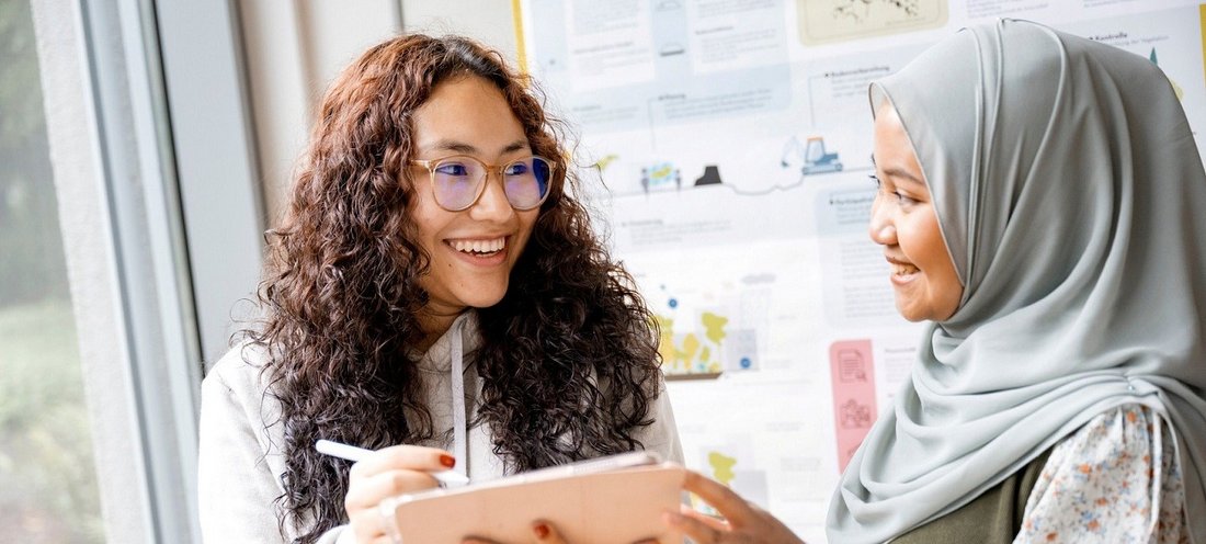Zwei internationale Studentinnen sprechen lächelnd über Inhalte auf einem Tablet.