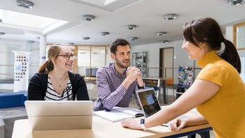 Drei Personen stehen an einem Tisch, mit aufgeklappten Laptops und Notizbuch und besprechen etwas