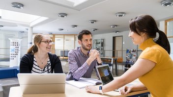 Drei Personen stehen an einem Tisch, mit aufgeklappten Laptops und Notizbuch und besprechen etwas