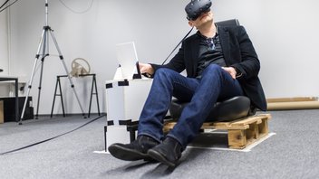 Ein Mann sitzt auf einem Autositz, angebracht auf einer Europalette und trägt einer VR-Brille, neben ihm steht ein Tablet