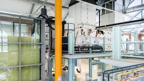 Eine Professorin steht mit drei Studierenden in einem großen Labor und erklärt eine Maschine