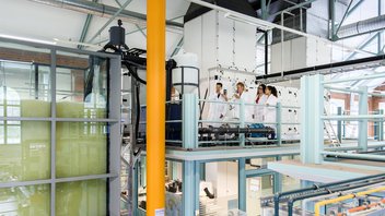 Eine Professorin steht mit drei Studierenden in einem großen Labor und erklärt eine Maschine