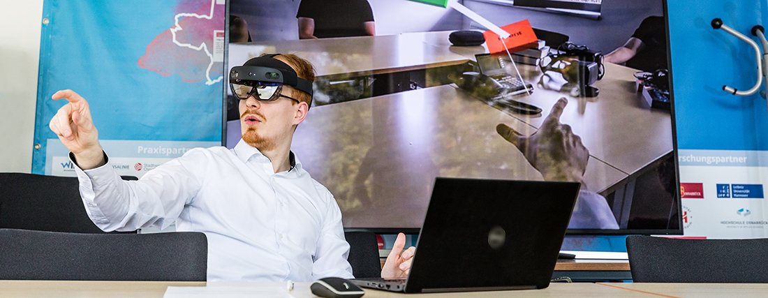 Ein Mann sitzt vor einem Laptop, er trägt eine AR-Brille und "tippt" mit dem Zeigefinger in der virtuellen Welt etwas an oder zeigt auf etwas. Im Hintergrund ist ein Meetingraum auf einer Leinwand projeziert, man erkennt darüber das, was der Mann durch die AR-Brille sieht